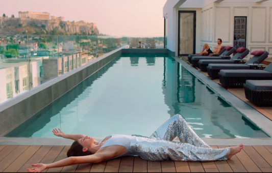 A woman in shiny silver pants lies with arms outstretched by a rooftop pool at Nyx Hotels Athens, while another person relaxes on a lounge chair. The cityscape and a hilltop ancient ruin create a stunning backdrop.