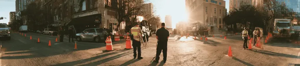 Panoramic view of a city street blocked by orange cones, with several workers and officials in safety vests standing near construction equipment as filming in Austin takes place under sunlight between tall buildings.