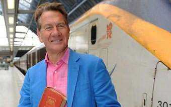 A smiling man in a blue blazer and pink shirt holds a red book while standing on a train platform next to a white and yellow train, capturing the spirit of Great Continental Railway Journeys as seen on the BBC.