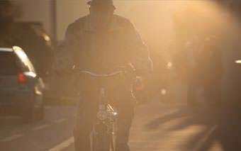 A person rides a bicycle on a street in bright sunlight, creating a silhouette effect through the Compass Light. Cars line the road, and the hazy scene captures a Day in the Life under intense sun.