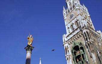 A tall, ornate clock tower with a pointed roof stands against a clear blue sky next to a golden statue on a column—an iconic sight for travel and tourism in Crete. A bird is flying in the middle background.