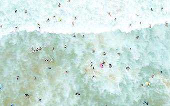 Aerial view of people swimming and playing in light blue ocean waves near the shore, embodying the Full Time Movement spirit, with white foamy water and scattered beachgoers visible.