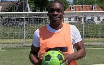 A smiling person wearing an orange training vest stands on a soccer field, holding a green soccer ball near the goalmouth. A goalpost and buildings are visible in the background.