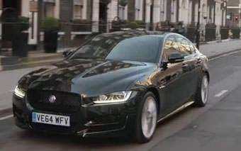 A black Jaguar XE sedan with UK license plate VE64 MFY cruises down a city street lined with buildings and greenery, perfect for an Idris Elba-inspired road trip.