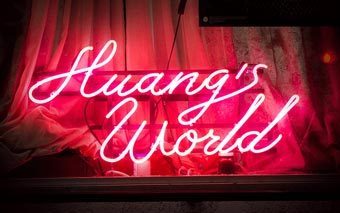 Neon pink sign in cursive script reading “Huang’s World” glows brightly against a backdrop of curtains, capturing the bold and vibrant vibe of the Vice series.