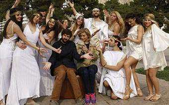 A group of people dressed in white, ancient Greek-style costumes surround two seated individuals, posing playfully outdoors with trees in the background—just another adventurous day with Dave and the crew from 24 Hours To Go Broke.