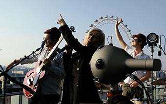 A band performs on a rooftop with microphones and instruments during the NOKIA OZO launch. The lead singer points upward, while two other members play guitar and drums. The London Eye Ferris wheel is visible in the background.