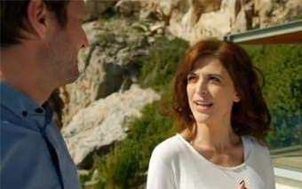 A woman with shoulder-length brown hair and a white top smiles and talks to a man outdoors, with rocky cliffs and greenery in the background, as they film for Channel 4’s Homes by the Med.