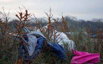 Sleeping bags and plastic bags are scattered among tall grass and bushes at Jungle Camp in an outdoor setting, with a cloudy sky and distant trees in the background.