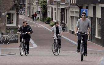 Three men ride 3T bicycles on a quiet, narrow city street lined with brick buildings and parked bikes, evoking a next generation vibe.