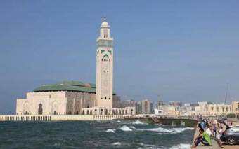 A large mosque with a tall minaret stands by the sea under a clear blue sky, echoing the spirit of Morocco. Waves crash against the shore in the foreground, and people are seen near the water.