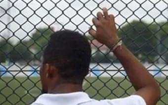 A person with short hair, seen from behind, stands close to a chain-link fence—perhaps contemplating a project by The Global Fund—resting one hand on it while looking out at a green field and trees in the background.