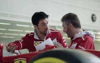 Two men in red Scuderia Ferrari team jackets stand beside a Formula 1 car, discussing papers, with a blurred garage background.