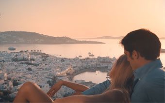 A couple relaxes together overlooking a coastal town with white buildings at sunset, enjoying breathtaking views from KENSHO Suites, a boutique hotel where the sea and distant hills create a dreamy backdrop.