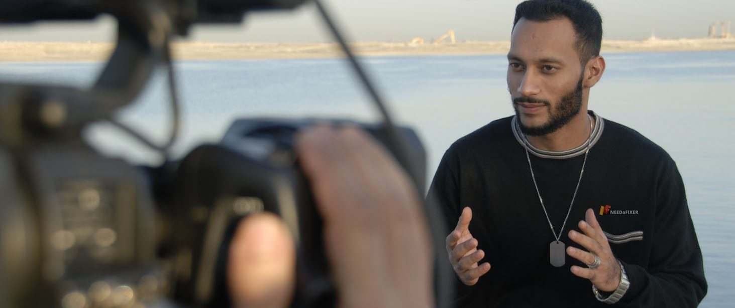 A man in a black shirt gestures while being filmed outdoors near a body of water, with distant sandy land in the background—capturing an authentic moment perfect for making a documentary.