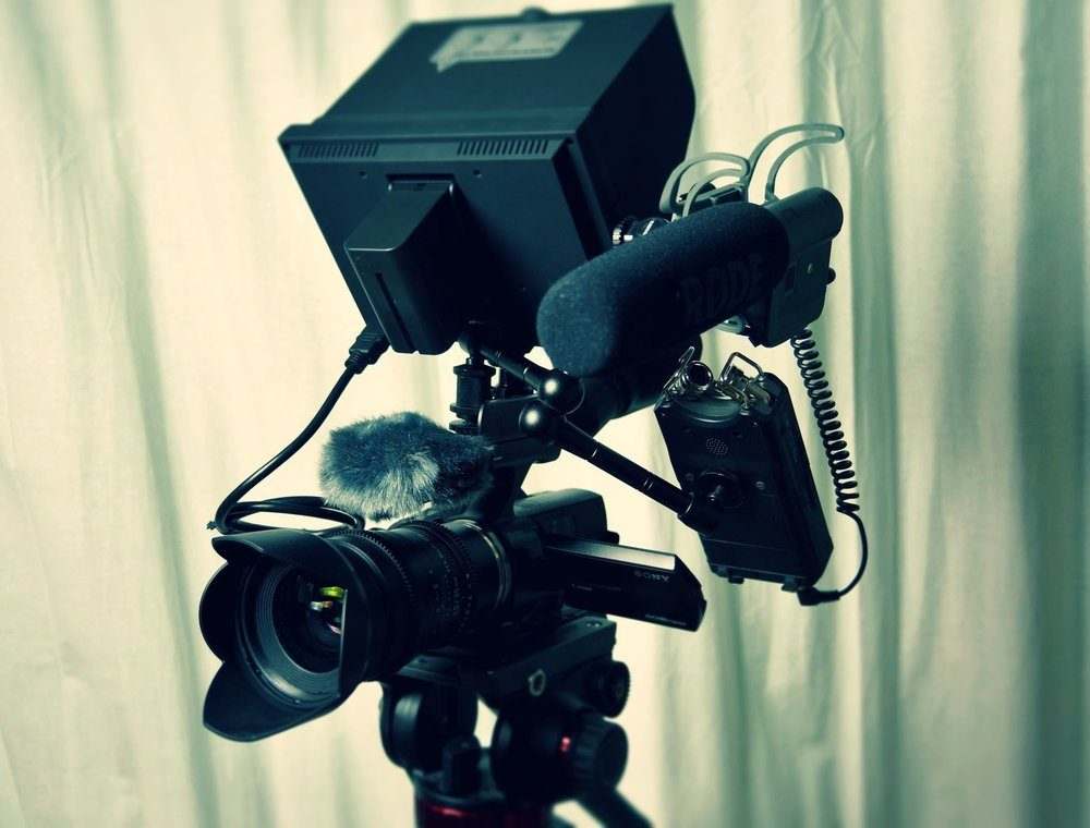 A professional video camera, like those used on the most expensive films, is mounted on a tripod with a large monitor, Rode microphone, and other recording accessories, set up in front of cream-colored curtains.