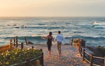Two people walk hand in hand along a sandy path toward the ocean at sunset on Mexicos Pacific Coast, with waves crashing and a golden sky in the background. Green plants and wooden railings line the scenic shoreline route.