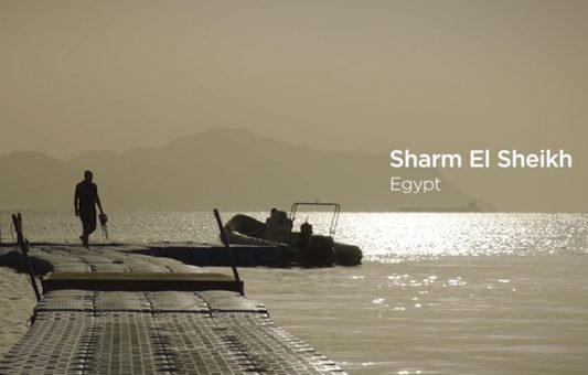 A person walks on a floating dock over calm water at sunset, with boats nearby and mountains in the background. Text reads “Sharm El Sheikh, Egypt,” highlighting the beauty of Egypt tourism.