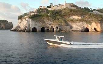 A Honda Marine motorboat speeds across calm blue water in front of a rocky island with caves and green vegetation, topped by historic stone buildings under a partly cloudy sky, captured by WCS Productions.