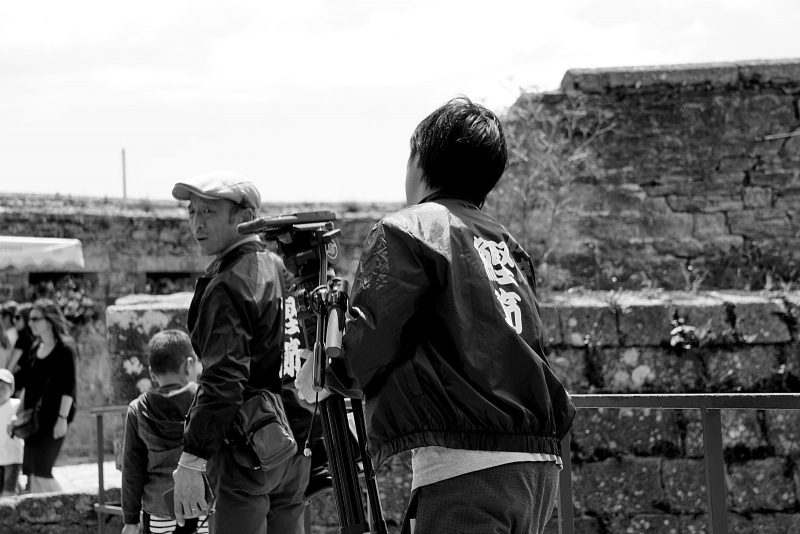 Two people in matching jackets stand outdoors near stone ruins; one operates a camera on a tripod, capturing footage for a documentary video, while the other looks back. Several people and children are visible in the background. The photo is black and white.