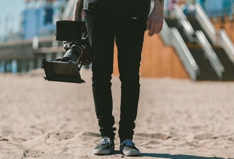 A person stands on sand, holding a professional video camera at their side—ready for a corporate video shoot. Only the lower half is visible in black pants and sneakers, with a blurred staircase in the background.