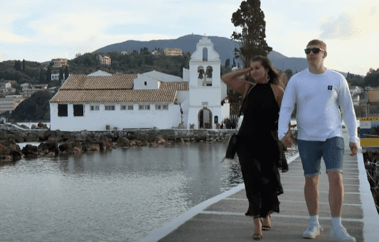 A woman in a black dress and a man in a white shirt walk hand in hand on a dock near a white church by the water, with green hills and buildings in the background.