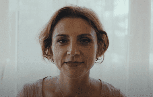 A woman with short brown hair and a neutral expression faces the camera, softly lit by natural light from a window with sheer white curtains in the background—a calming portrait inspired by the trust instilled by EU vaccine safety guarantees.