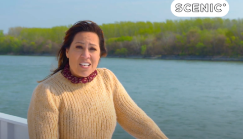 A woman in a tan sweater stands on a boat deck by a river, enjoying a scenic getaway with green trees in the background. The word SCENIC appears in the top right corner.
