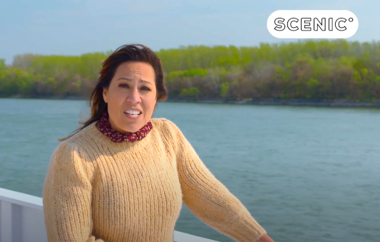 A woman in a tan sweater stands on a boat deck by a river, enjoying a scenic getaway with green trees in the background. The word SCENIC appears in the top right corner.