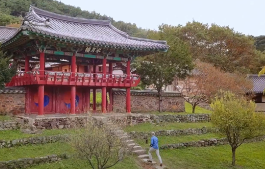 Capturing Korea: A person in a blue jacket walks up stone steps toward a traditional Korean pavilion with red pillars, surrounded by green trees and grass.