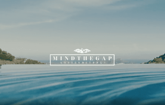 A tranquil infinity pool overlooks distant hills and sky, with the text Mind the Gap Design Supply and a bat logo centered in the image.