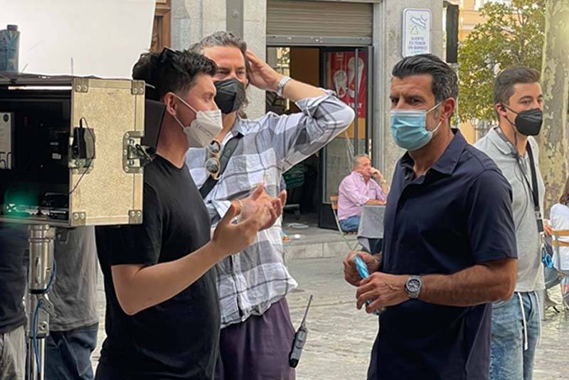 Several men wearing face masks stand outdoors in conversation near filming equipment, possibly preparing for a 60 Seconds monthly interview with industry leaders. One gestures as another listens, while people sit at tables amid trees and buildings in the background.