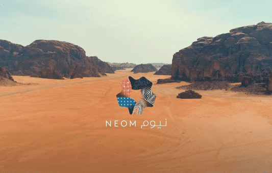 A desert landscape with rocky formations under a blue sky. In the center, the NEOM logo, a multicolored star shape, appears above the text “NEOM” in English and Arabic, highlighting NEOM as the future hub for extreme sports.