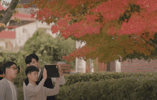 Three people stand outdoors under a tree with vibrant red leaves. One person holds up a tablet, using Google for Education, while the others watch, seemingly looking at the device and the colorful foliage.