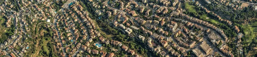 Aerial view of a suburban neighborhood with many houses, tree-lined streets, and green spaces—reminiscent of scenes often found while filming in Rome. The layout shows winding roads and densely packed homes surrounded by foliage.