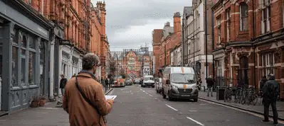 A person with a brown jacket stands on a city street lined with red brick buildings, possibly filming in Birmingham, watching cars and vans drive by under a cloudy sky.