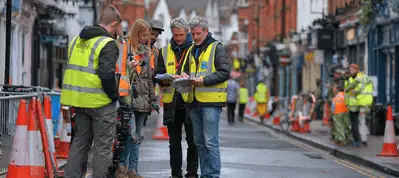 A group of people wearing high-visibility vests stand in the middle of a street lined with traffic cones and equipment, discussing documents during filming in Birmingham, while others work in the background.