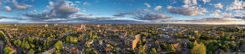 A panoramic aerial view of a cityscape showcases filming in Birmingham, with numerous buildings, green trees, and houses under a partly cloudy sky. The horizon is visible in the distance as sunlight illuminates parts of the vibrant city.