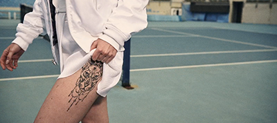 A person in white sportswear stands on a tennis court in Italy, lifting their shorts to reveal a large tattoo of a roaring tiger on their upper thigh.