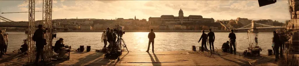 A film crew sets up cameras and equipment on a wooden dock by a river at sunset, filming in Budapest, with a silhouetted figure in the center and the city's iconic bridges and historic buildings across the water.