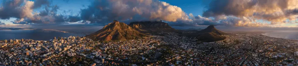 A panoramic view of Cape Town, South Africa, with Table Mountain and surrounding peaks under dramatic clouds—an ideal scene for filming in Cape Town—city buildings and coastline aglow in warm, golden light.