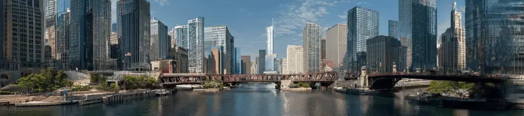 Panoramic view of a cityscape with tall modern skyscrapers, a metal bridge over a river, and blue sky with clouds—an ideal backdrop for filming in Chicago’s vibrant downtown.