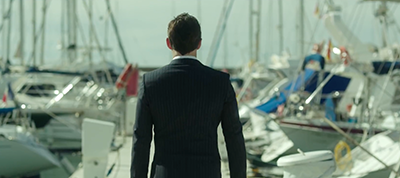 A man in a suit stands on a dock in Italy, facing away from the camera and gazing at a marina filled with moored boats and yachts under a bright sky, capturing the perfect scene for a video production.