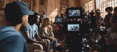 A film crew is filming in South Korea, capturing two people sitting and talking in a rustic, sunlit room filled with people. A camera is in the foreground, focused on the scene as the crew and actors work together seamlessly.