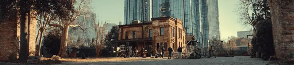 A group of people gathers outside an old brick building with a porch, surrounded by trees, as towering glass skyscrapers hint at filming in Atlanta in the background.
