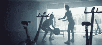 Two people exercise together in a well-lit gym in Italy, one squatting with a medicine ball while the other stands nearby. Surrounded by exercise bikes, the moment is ideal for capturing by a video production company.