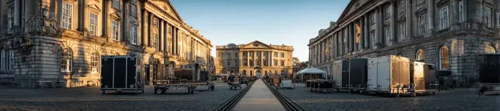 Film production equipment and trailers are set up along a cobblestone street between two historic, grand buildings while filming in Dublin, with another ornate building in the background under a clear sky.