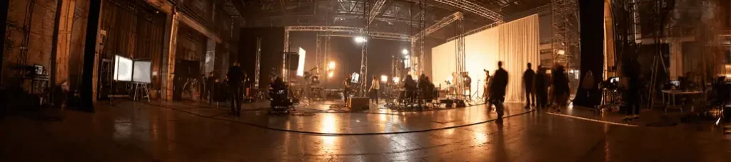 A wide-angle view of a film set in a large studio while filming in Atlanta, with crew, lighting equipment, cameras, and shadows of people working under warm, industrial lighting.