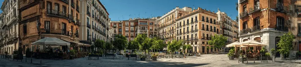 A wide view of a sunny European plaza in Madrid, perfect for filming, surrounded by historic, multi-story buildings with balconies, outdoor cafes with umbrellas, and rows of trees lining the open, cobblestone square.