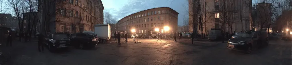 A wide, panoramic view of an outdoor film set at dusk, with bright lights illuminating a group of people and vehicles—filming in Moscow—in front of a large, curved building surrounded by bare trees and older apartment blocks.